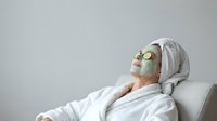 Woman relaxing in spa chair wearing green facial mask and white bathrobe with towel on head for skincare treatment and wellness routine.
