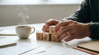 Senior businessman hands arranging wooden blocks with business icons on office desk with steaming coffee cup and documents for strategic planning.
