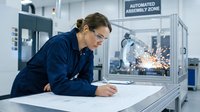 Engineer in blue uniform reviewing technical blueprints at workstation with automated assembly machinery and robotic welding in background.