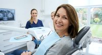 Middle-aged Caucasian woman smiling in dental chair during checkup with female dentist and assistant in modern clinic office for oral health care.