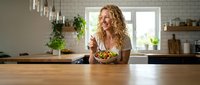 Young blonde woman enjoying healthy salad meal in modern kitchen with wooden countertop and pendant lighting. Lifestyle nutrition wellness concept.