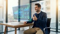 Professional businessman celebrating success with thumbs up gesture while viewing financial charts and data analytics on computer screen in modern office.