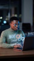 Young African American software developer working on laptop with holographic data visualization interface in modern office at night.