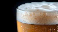 Close up view of fresh beer with thick white foam head and bubbles rising in amber liquid against dark background, macro photography of beverage.