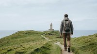 Male hiker with backpack walking on coastal trail toward lighthouse on grassy hillside, adventure and outdoor exploration concept for travel and nature themes.