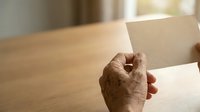 Senior adult hand holding blank cream envelope on wooden table with natural window light, suitable for mail, correspondence, and business communication concepts.