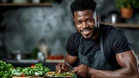 Young African American male chef smiling while preparing fresh salad in modern kitchen wearing black apron for culinary cooking and food preparation.