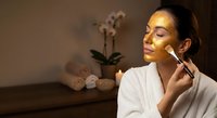 Young Hispanic woman in white bathrobe applying skincare serum with dropper in luxury spa setting with orchid flowers and towels for wellness routine.