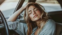 Young blonde woman experiencing headache or fatigue while sitting in car interior, holding head with hand in stress or exhaustion gesture.