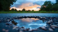 Dramatic sunset sky reflected in puddle on rough asphalt road with stones and pebbles around edges, moody nature landscape with forest silhouette background.