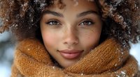 Young African American woman with natural curly hair and freckles wearing cozy orange scarf in winter snow, close up portrait showing genuine smile and bright blue eyes.