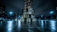 Modern stacked concrete tower sculpture in urban plaza at night with wet pavement, city lights, and contemporary architecture for design projects.