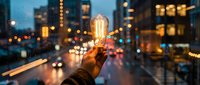 Hand holding glowing vintage Edison bulb against blurred city lights at dusk. Creative inspiration and innovation concept for business presentations and marketing.