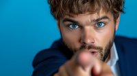 Young Caucasian businessman with blue eyes and beard pointing finger at camera in confident gesture against bright blue background for marketing campaigns.