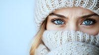 Close up portrait of bright blue eyes and blonde hair peeking through white knitted winter hat and cozy scarf wrapped around face against light background.