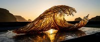 Stunning golden wave sculpture made of intertwined branches creates dramatic silhouette against ocean sunset backdrop with mountains and reflective wet sand beach.
