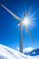 Wind turbine against bright sun and blue sky on snowy mountain peak. Clean energy generation in winter alpine landscape with sunburst effect.