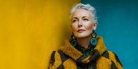Elegant mature woman with blonde hair wearing yellow coat and turquoise earrings against vibrant yellow and teal background.