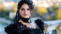 Young woman in elegant black Victorian dress with lace details and dark hair styled in romantic updo poses outdoors against blurred natural background.
