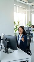 Young South Asian woman in business casual attire giving thumbs up at desk with computer in bright modern office with colleagues and plants.