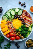 Fresh bowl with sliced cucumber, cheddar cheese, quinoa, cherry tomatoes, broccoli, olives, bacon and mango arranged in colorful sections on white plate. Mediterranean style lunch.
