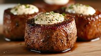 Juicy grilled beef tenderloin medallions topped with herb butter and black pepper on wooden cutting board, close-up view of gourmet steak preparation.