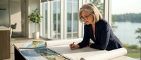 Professional mature Caucasian businesswoman architect reviewing building blueprints at modern office desk near large windows with natural lighting.