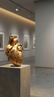 Golden anatomical heart sculpture displayed on pedestal in modern art gallery with contemporary lighting and framed artworks on walls.