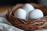 Fresh white eggs in rustic woven basket on light linen cloth, macro food photography with soft natural lighting and shallow depth of field.