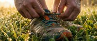 Male hiker tying hiking boot laces on grass during golden hour outdoor adventure. Closeup of hands securing footwear before trekking.