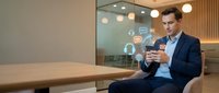 Businessman in dark suit holding smartphone with digital icons floating on screen in modern office workspace with glass partition and warm lighting.