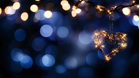 Glowing heart-shaped decoration with warm fairy lights hanging against dark blue bokeh background creates romantic festive atmosphere for Valentine's Day or wedding design.