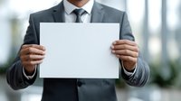 Businessman in gray suit holding blank white paper at office space, closeup view of male hands presenting empty mockup sheet for business message.