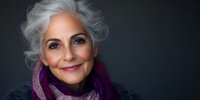 Elegant mature woman with silver hair and warm smile wearing purple scarf against dark background, radiating confidence and natural beauty in professional portrait.