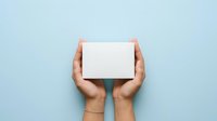 Hands holding blank white box against light blue background, suitable for product packaging mockups, gift presentations, and commercial advertising campaigns.