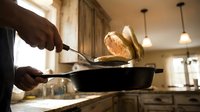 Professional chef flipping golden pancakes in cast iron skillet during breakfast preparation in modern kitchen with warm lighting and wooden cabinetry.