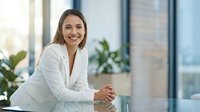 Professional businesswoman in white blazer smiling confidently at modern office desk with city view background for corporate presentations.