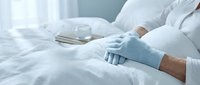 Healthcare professional in protective gloves preparing medical bed with clean white linens in sterile hospital environment for patient care procedures.