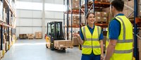Warehouse workers in safety vests discussing logistics operations with forklift and storage shelves in industrial distribution center background.