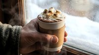 Hot chocolate with marshmallows and cinnamon in takeaway cup held by hand near snowy window. Cozy winter drink for cold weather comfort and warmth.