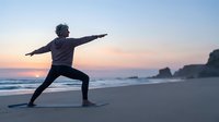 Young Asian man practicing warrior yoga pose on beach at sunset with ocean waves and peaceful sky for wellness and meditation content.