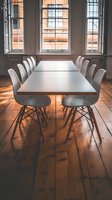 Modern conference room with white chairs and long table, sunlight streaming through tall windows onto wooden floors. Natural lighting creates dramatic shadows.