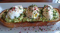 Toasted bread topped with mashed avocado and three poached eggs, garnished with fresh herbs and red pepper flakes, served on rustic wooden surface.