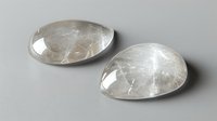 Polished clear quartz crystal pear shaped cabochons on light gray background, natural gemstone pair with translucent rutile inclusions for jewelry making.