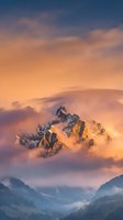 Dramatic mountain peak emerging through golden clouds at sunrise with warm orange and pink sky colors creating majestic alpine landscape scenery.