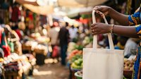 Shopper holding empty canvas tote bag at bustling outdoor market with fresh produce and vendors in background, sustainable shopping and eco-friendly lifestyle concept.