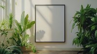 Empty black frame mockup on white wall surrounded by green houseplants with natural sunlight and shadows creating minimalist home interior design scene.
