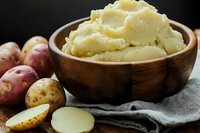 Creamy mashed potatoes served in wooden bowl with raw red potatoes on rustic wooden surface, close up view of traditional comfort food preparation.