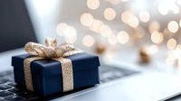 Navy blue gift box with sparkling gold ribbon on laptop keyboard against soft bokeh lights background, festive holiday shopping and online gifting concept.