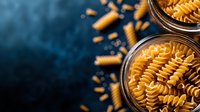 Raw fusilli pasta in glass jars on dark blue background with scattered dry noodles. Minimalist food composition with dramatic lighting and copy space.
